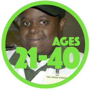 Close up of a smiling young man of color with a developmental disability who is wearing a work uniform with green cap and white polo shirt with green color. Name tag says, “The Fresh Market.”