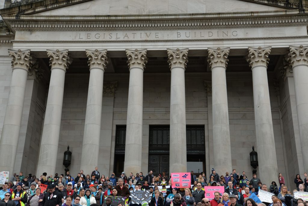 Front of Legislative Building in Olympia. Crowd of disability advocates waving signs on the front steps.