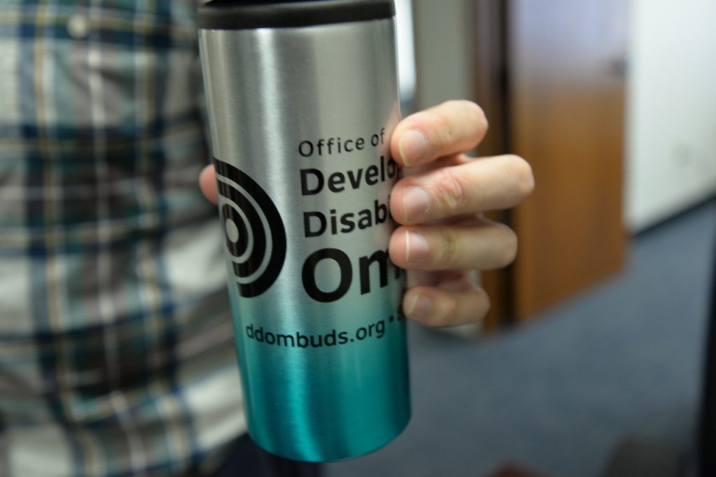 Hand holding a travel mug with Office of Developmental Disabilities logo and website url ddombuds.org