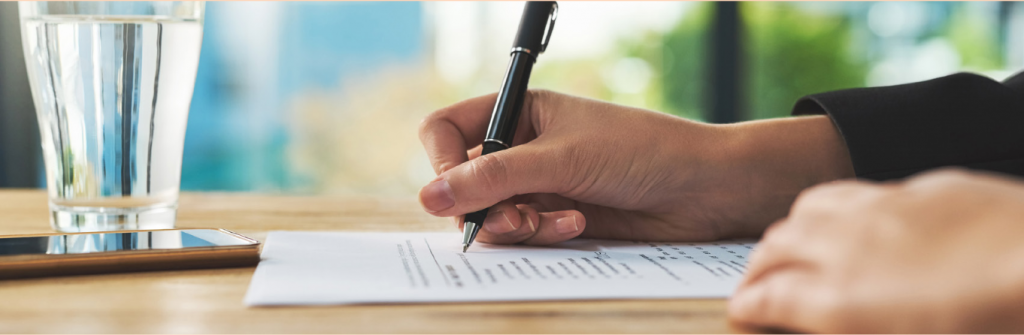 Side view of a woman's hand holding a pen over a piece of paper. Window in the background and a glass of water on the table.