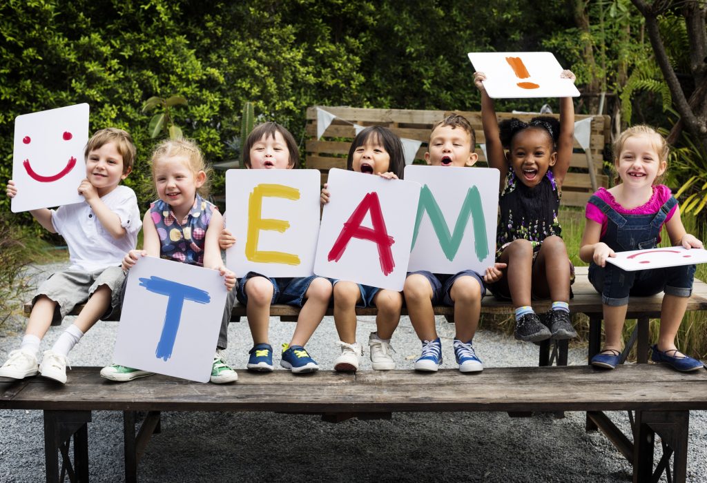 Children are sitting on the wooden table, each holding a different colored letter of the word "team."