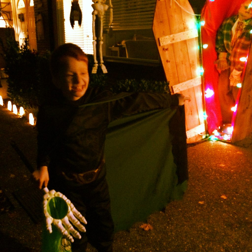 Nate outside on Halloween night, dressed in black and holding a skelton hand, grinning. In the background, a casket stands upright with a zombie inside.