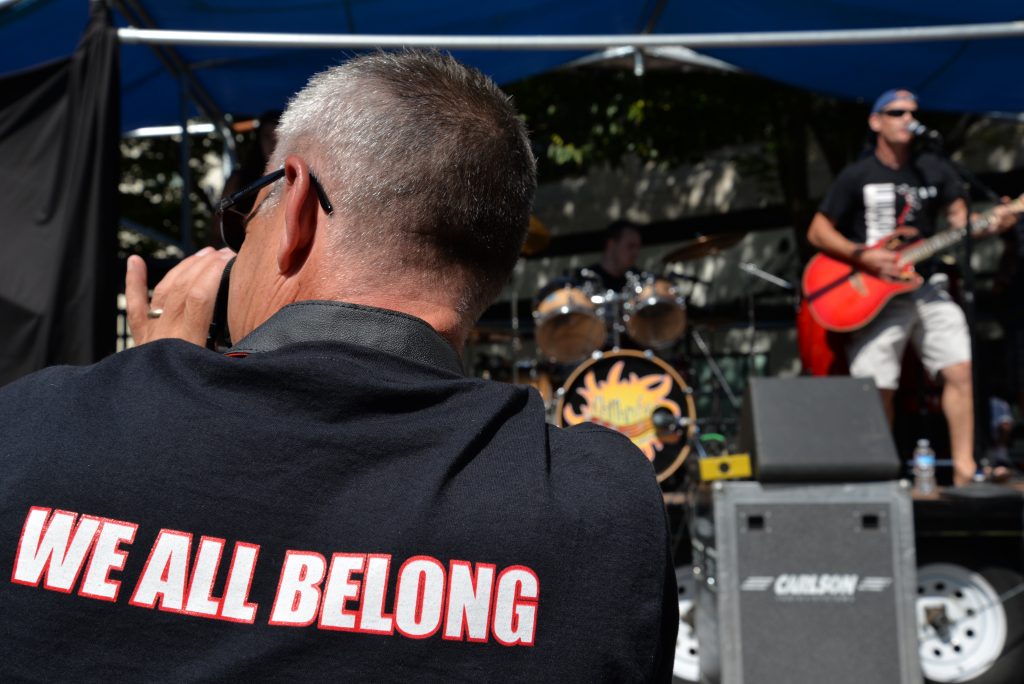 Outdoor festival, band playing in background. Forergound image of the back of a man wearing a t-shirt with the words We All Belong.