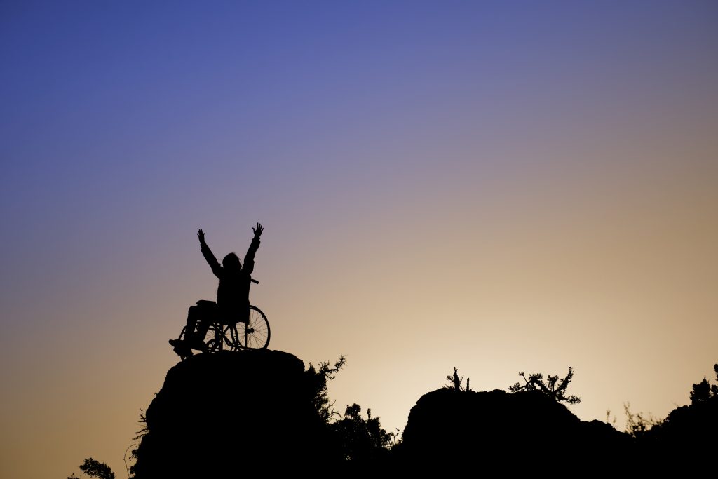 Silhouette of girl in a wheelchair on a rock outcropping, arms reaching toward the sky.