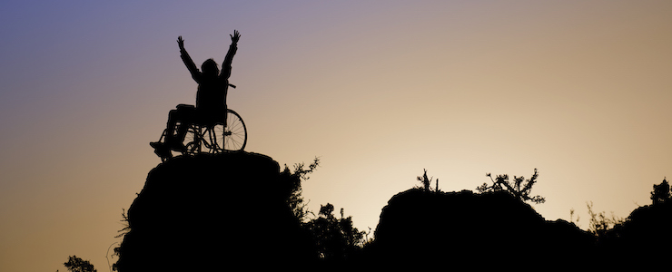 Silhouette of girl in a wheelchair on a rock outcropping, arms reaching toward the sky.