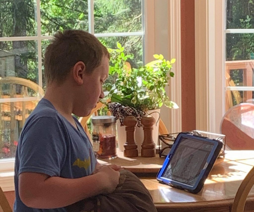 Nathan at home, sitting at a table with iPad during speech therapy.