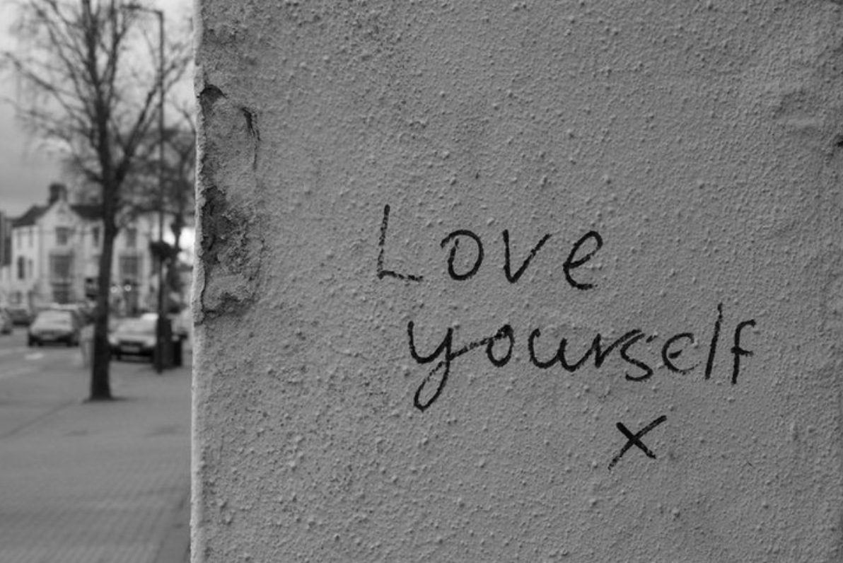 Exterior lack and white photo of a concrete wall with the handwritten words, Love yourself x. Small town in the background.