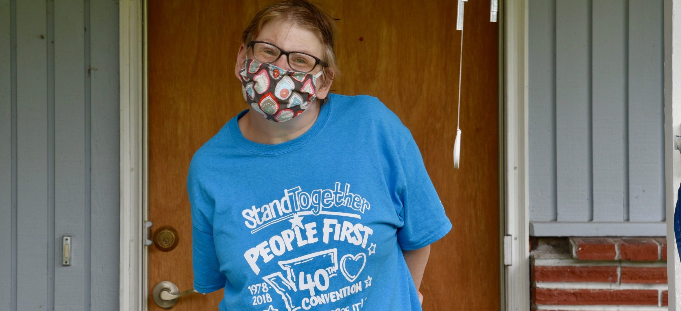 Disabled woman with People First t-shirt wearing a mask, standing outside on a porch.
